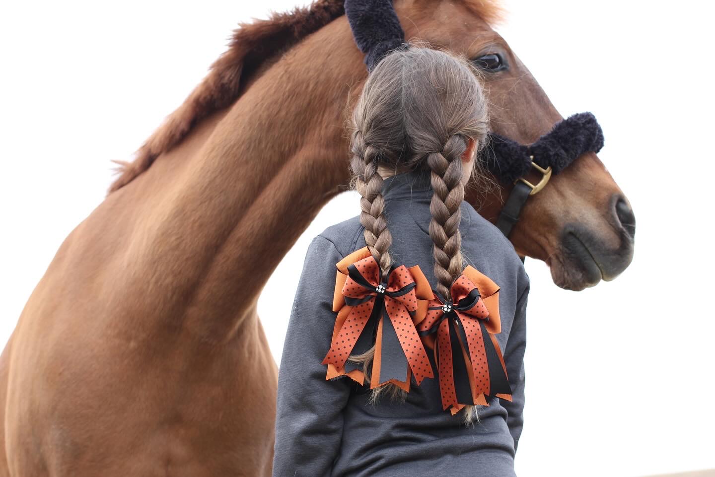 A young rider with braided hair wearing orange and black polka-dot Best in Bow bows, with her chestnut pony resting beside her